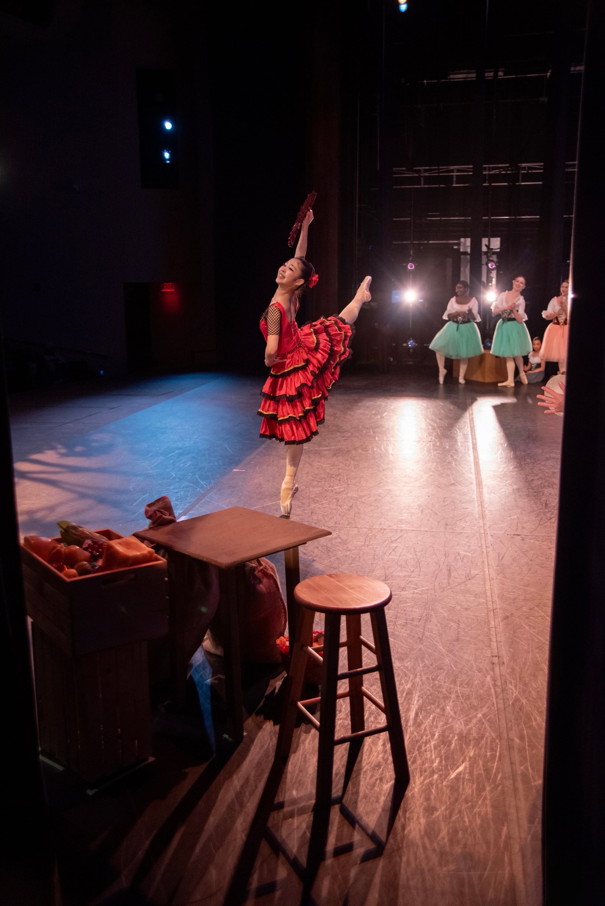 Kitri mid-leap in a red ruffled dress, seen past a dancer in the wings